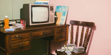 Smells Every Gen-X Person Will Remember From Their Slightly Toxic Childhood A retro 1990s basement setup with a classic CRT TV, a gray gaming console on the floor with controllers, loose game cartridges, and VHS tapes on a shelf against a wall.