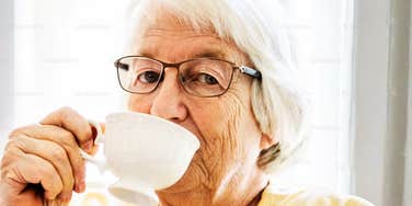 A close-up portrait of a thoughtful senior citizen enjoying a hot beverage inside a bright, clean room; illustrating 'the intergenerational quality fracture' where older and younger cohorts share identical frustrations with modern tech and corporate systems.