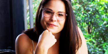 A close-up portrait of a woman with glasses smiling warmly outdoors in soft, natural light; a visual representation of 'the compassionate person