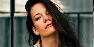 A candid outdoor street portrait of a fashionable young woman wearing a colorful printed top, looking directly into the camera with an intriguing, calm expression.