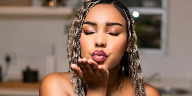 A woman leaning forward in a bright, minimalist white kitchen while blowing an exaggerated kiss to the camera; a visual representation of 'the overly staged persona' archetype used to study validation-seeking behavior.