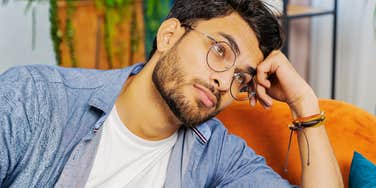 A young Indian man resting his chin on his hands while sitting on a couch, looking up thoughtfully with a stressed, weary expression.