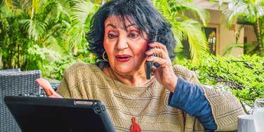 An older woman relaxing in outdoor sunlight, attentively using a smartphone and tablet, illustrating modern tech adoption and daily AI interaction.