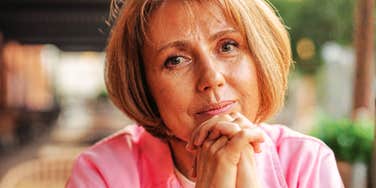 A mature woman wearing a pink shirt resting her hands under her chin with a contemplative expression, symbolizing generational differences, cultural shifts, and communication dynamics.