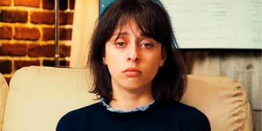 A young woman with dark hair sitting indoors with dark under-eye circles and a glum face, symbolizing burnout, chronic misery, and emotional fatigue.