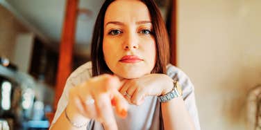 A close-up portrait of a woman resting her chin on her hands while pointing her finger directly into the camera with a serious, intense expression.