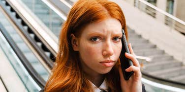 A close-up portrait of a young woman with long red hair and freckles holding a smartphone to her ear with an annoyed, tired expression near an escalator.