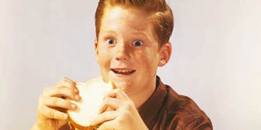 A child enjoying a sandwich indoors, symbolizing 1970s domestic life, retro lunchbox culture, and generational food preferences.