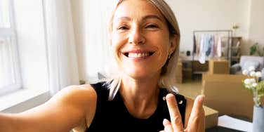 A happy middle-aged woman taking a cheerful selfie with her smartphone while sitting at her office workspace.