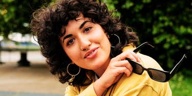 A fashionable young woman with curly hair relaxing outdoors with sunglasses, symbolizing modern culture, Gen-Z communication, and confident self-expression.