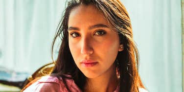 A close-up portrait of a young woman with dark hair wearing a pink top looking directly into the camera with a guarded, serious expression.