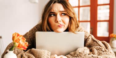 A thoughtful young woman holding a open laptop while looking off-camera with a contemplative expression.