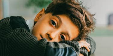 A young boy wearing a long-sleeved sweater resting his head on a desk while looking directly into the camera with a thoughtful gaze.
