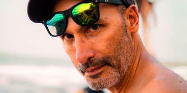 A close-up portrait of a mature man wearing a baseball cap peering over green reflective sunglasses outdoors.