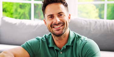 A warm lifestyle portrait of a smiling mid-adult man relaxing comfortably at home on a couch, looking approachable and content.
