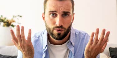 A young man with a beard making a confused gesture while sitting in a domestic setting, symbolizing communication breakdowns, low emotional intelligence, and relationship frustration.