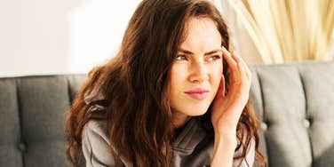 A young woman with long dark hair sitting on a couch, squining in discomfort and holding her hand to her temple.