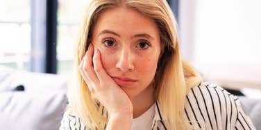 A young adult woman wearing a striped shirt sitting on a home couch in deep reflection, symbolizing introspection, emotional processing, and generational boundaries.