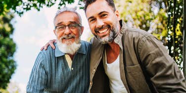 Father and his adult son smile warmly into the camera with arms around each other 