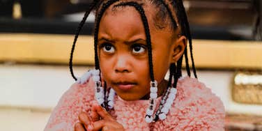 A close-up portrait of a young Black girl with braided hair and white beads wearing a pink fuzzy sweater, looking up thoughtfully with a serious expression.
