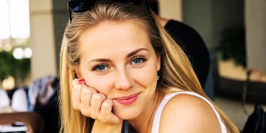 A young woman smiling thoughtfully while relaxing at a cafe table, symbolizing active listening, verbal presence, and intellectual charm.
