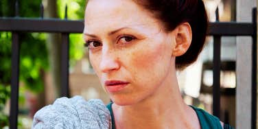 A close-up portrait of a red-haired woman with freckles looking directly at the camera with a stoic, contemplative, and somber expression.