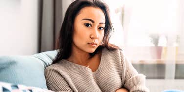 A puzzled and upset young woman sitting on a sofa with her arms crossed, looking troubled in a living room setting.