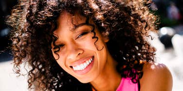A young woman with voluminous curly hair laughing outdoor in bright sunlight, symbolizing warmth, emotional maturity, and natural social class.