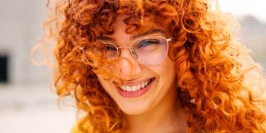 A close-up portrait of a cheerful redhead woman with long curly hair and glasses, smiling warmly into the camera.
