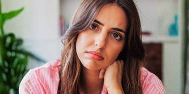A young woman wearing a pink striped shirt leaning on her hand with a contemplative, guarded expression, symbolizing social manipulation, deceptive cues, and psychological self-defense.
