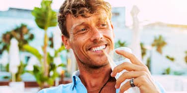 A close-up portrait of a handsome man with blond hair sipping a drink through a straw on a sunny hotel terrace.