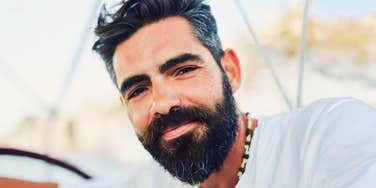 A close-up portrait of a handsome bearded man with dark hair, looking directly into the camera with a calm, relaxed, and cheerful expression.