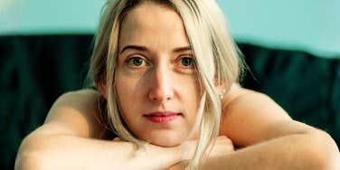 A close-up portrait of a woman resting her head gently on her arms with a reflective, quiet expression.
