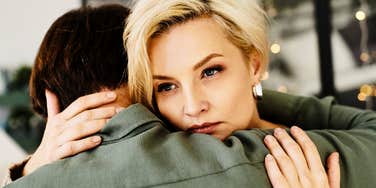 A blonde woman holding her partner close indoors with a conflicted and sad expression, symbolizing emotional distance, relationship missteps, and quiet doubt.