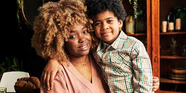 A warm portrait of a Black woman with curly blonde-highlighted hair embracing her young son indoors while smiling at the camera.