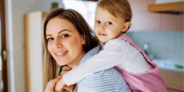 A smiling mother in a striped shirt giving her young toddler daughter a piggyback ride at home.