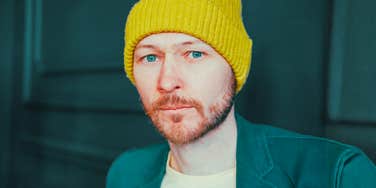 A close-up portrait of a bearded man in a yellow beanie and green jacket looking suspicious and skeptical against a dark blue background.