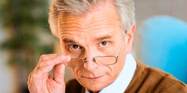 An older man looking directly into the camera while lowering his eyeglasses, symbolizing curiosity, sharp observation, and generational perspective.