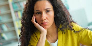 A pensive woman sitting indoors with her hands clasped near her face, looking upward in deep thought and self-reflection.