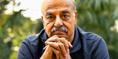 A close-up portrait of a senior Indian man with a mustache resting his chin on clasped hands, looking forward with a pensive, somber expression outdoors.