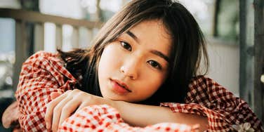 A close-up portrait of a young East Asian woman with straight dark hair wearing a red checkered shirt, leaning her head on her arms outdoors.