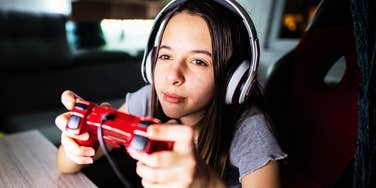 My Twelve-Year-Old Was Asked to Show Her Body to Play PlayStation A candid portrait of a young girl sitting in a living room, holding a wireless gaming controller and gazing intently at a glowing screen.