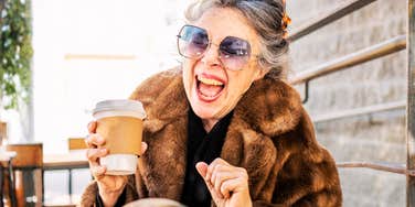 A candid portrait of an expressive senior woman wearing big sunglasses, a fur coat, and hair clips, laughing exuberantly while holding a coffee cup.