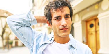 A man in a light blue denim shirt scratching his head outdoors, symbolizing uncertainty, awkward speech patterns, and deep reflection.