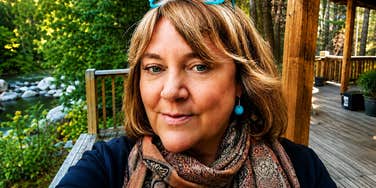 A selfie portrait of a mature woman with blonde hair, bright blue glasses resting on her head, blue drop earrings, and a patterned scarf, standing outdoors on a wooden deck near a river.
