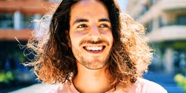 A man with long curly brown hair and a light beard smiling brightly in an outdoor urban setting, symbolizing mental clarity, happiness, and freedom from screen fatigue.