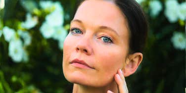 A close-up portrait of a woman with light blue eyes and dark hair resting her hand on her cheek against a background of white flowers.