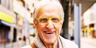 A senior man wearing bright green reading glasses and a striped scarf grinning happily on a city street, symbolizing contentment, life experience, and practical wisdom.
