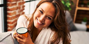 Smiling woman holds coffee mug on couch, embodying the warmth and self-awareness of people who handle forgetting a name gracefully.