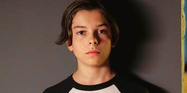 A young boy standing indoors with dramatic lighting casting shadows behind him, looking somber and thoughtful, symbolizing childhood trauma, quiet grief, and emotional resilience.